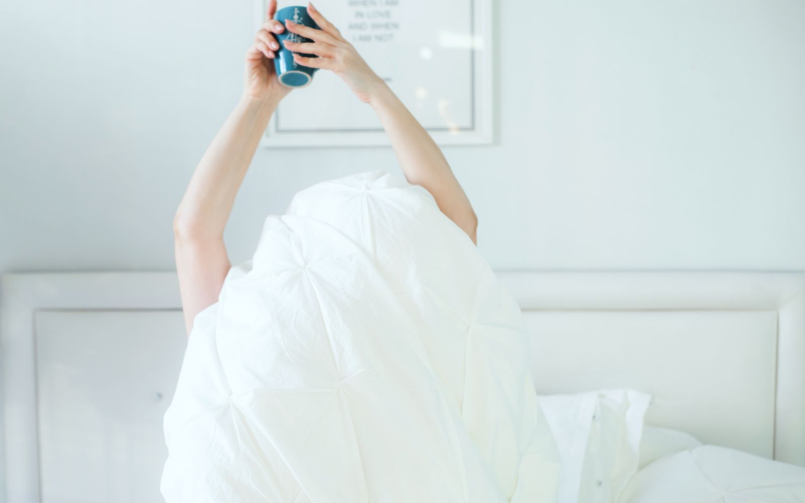 A person holding a coffee mug while relaxing in bed, covered with a white sheet.