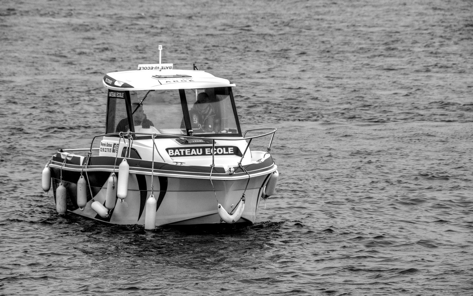 A black and white image showing a training boat labeled 'Bateau Ecole' on a calm sea.